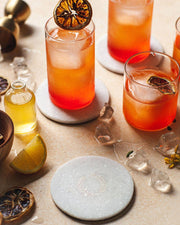 Farmhouse Pottery - Marble Laurel Coasters elegantly displayed with cocktails, showcasing their protective function amidst a decorative setup of glassware and citrus garnishes.