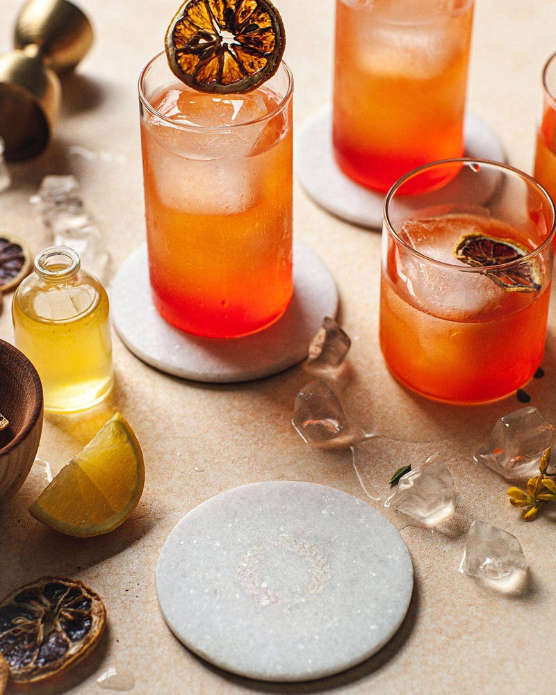 Farmhouse Pottery - Marble Laurel Coasters elegantly displayed with cocktails, showcasing their protective function amidst a decorative setup of glassware and citrus garnishes.