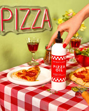 Brightland Pizza Oil bottle amidst a pizza setup, with slices and a hand reaching for it, highlighting its use for enhancing flavors.