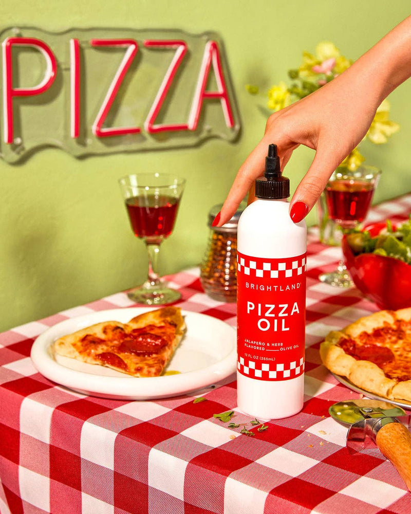 Brightland Pizza Oil bottle amidst a pizza setup, with slices and a hand reaching for it, highlighting its use for enhancing flavors.