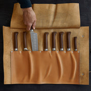 Opinel | 8 Slot Chef Knife Carrier, Leather, showcasing a hand lifting a Santoku knife, with eight wooden-handled knives neatly stored in a tan leather roll.