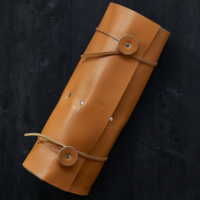 Opinel | 8 Slot Chef Knife Carrier, Leather: A tan leather roll-up pouch designed for storing chef knives, featuring stitched straps and snap buttons on a dark wooden surface.
