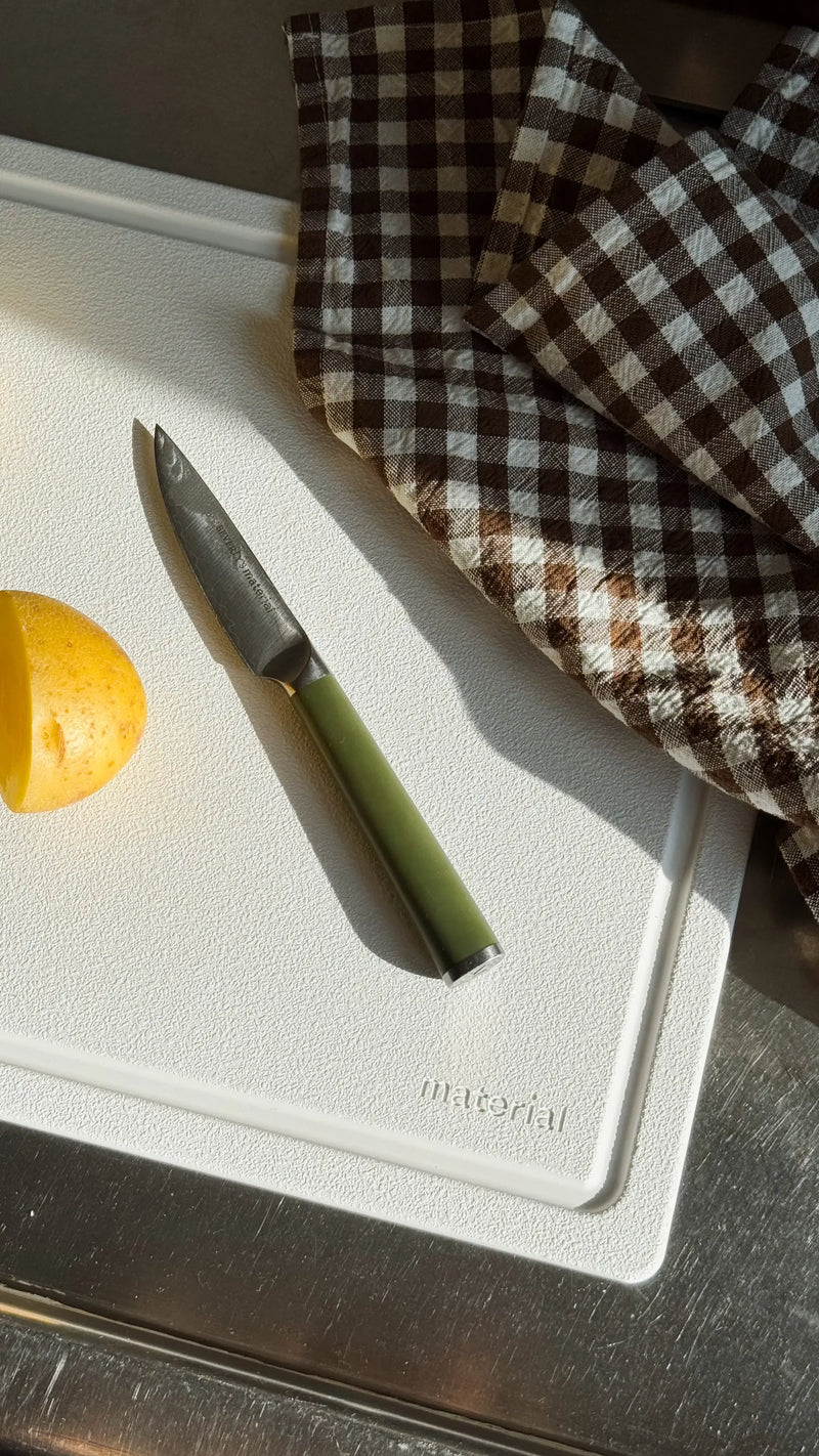 Material - The MK Free Board: Cloud, featuring a paring knife, potato, and checkered cloth, showcased on a stainless-steel surface for efficient, eco-friendly kitchen prep.