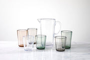 Zwiesel Fortessa | Veranda Double Old Fashioned Clear 14oz Tumbler with a clear pitcher, surrounded by six subtly tinted tumblers on a light surface.