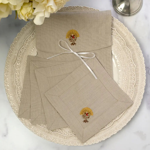 Arte Italica & Crown Linen Designs Turkey Cocktail Napkins on a charger, featuring embroidered sheaf-of-wheat motif, tied with ribbon; roses and salt shaker in background.