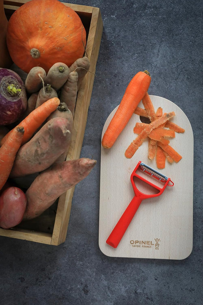 Opinel | T-Duo Peeler, Green, on a board with a peeled carrot and skins, near a crate of assorted root vegetables, showcasing versatile kitchen tool functionality.