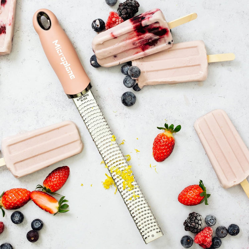 Microplane | Premium Zester, Grater - Dusty Rose with lemon zest, surrounded by berry-swirl ice pops and fresh berries on a countertop.