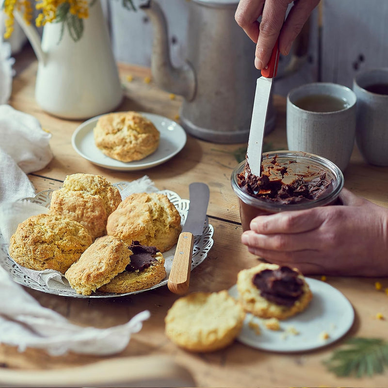 Opinel | Brunch Knives, Natural: Hands using the knife to scoop spread from a jar, surrounded by scones and mugs on a rustic kitchen table.