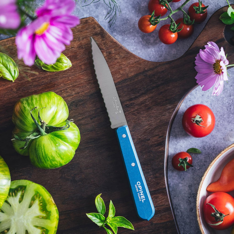 Opinel | Essential Serrated Knife, Skyblue, on a cutting board with heirloom tomatoes, cherry tomatoes, basil, and cosmos flowers in a colorful prep scene.