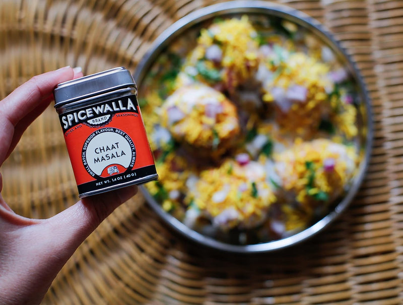 Hand holding Spicewalla Chaat Masala tin from the Chai Pani Masala Collection 6 Pack, with Indian street-food bowl in the background.