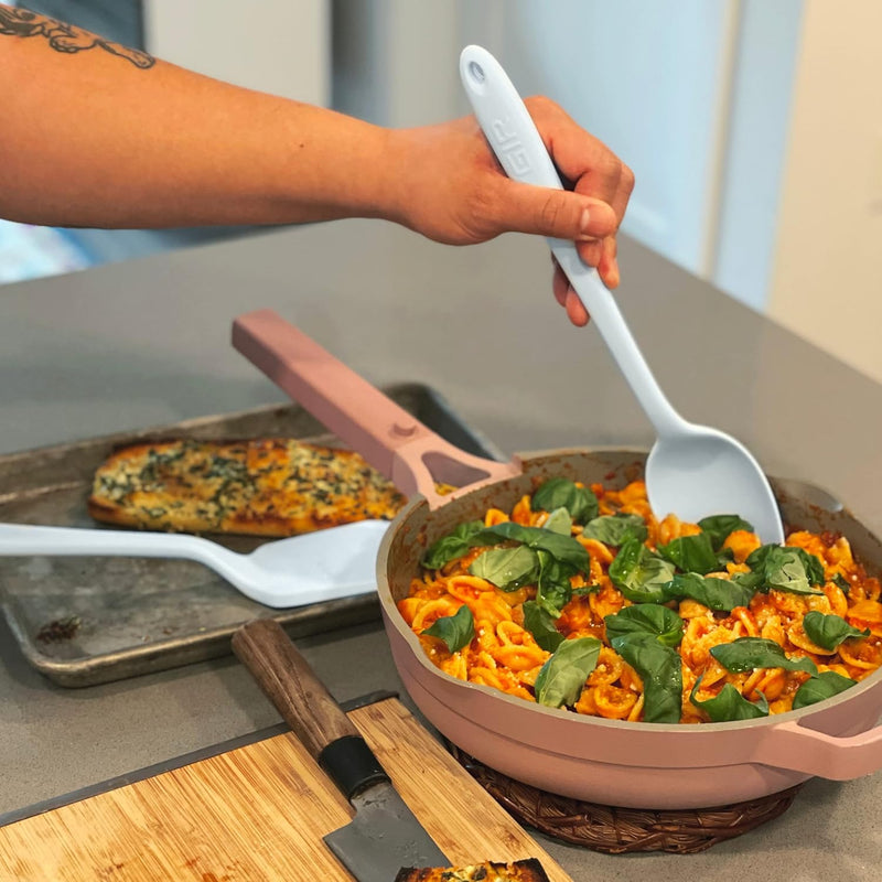 GIR | Ultimate Spoon - Studio White: A person uses the versatile kitchen tool to stir food in a pan, highlighting its deep bowl and flexible edges.