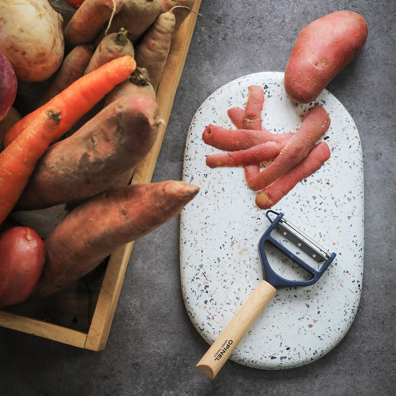 Opinel | T Duo Peeler with Wooden Handle, Blue, showcased on a terrazzo board, surrounded by carrots and potatoes, features dual blades for peeling and julienning.