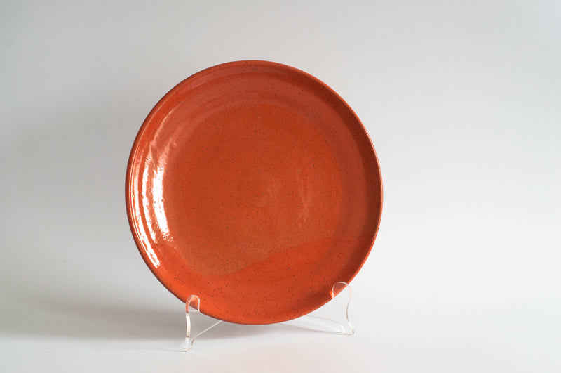 RachaelPots - Salad Plate: Coral, a hand-thrown, glossy orange-red ceramic plate with dark speckles, displayed on an easel, ideal for brightening your table setting.