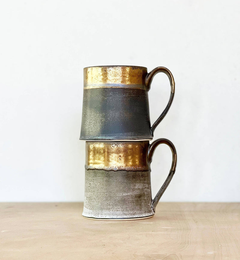 Two stacked Jessica Wertz Ceramics - Black + Gold Mugs with metallic-gold rims and matte grayish bodies on a wooden surface. Each mug holds 12 ounces and is dishwasher safe.