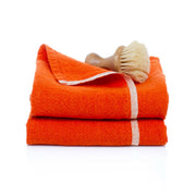 Caravan Home | Chunky Linen Orange Towels feature bright-orange linen with a white border, stacked with a wooden-handled brush, offering a modern rustic aesthetic.