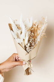Idlewild Floral Co. - Snow Dried Flower Bouquet: A hand holds a small, beige-toned bouquet of dried flowers and grasses, wrapped in kraft paper and tied with twine.