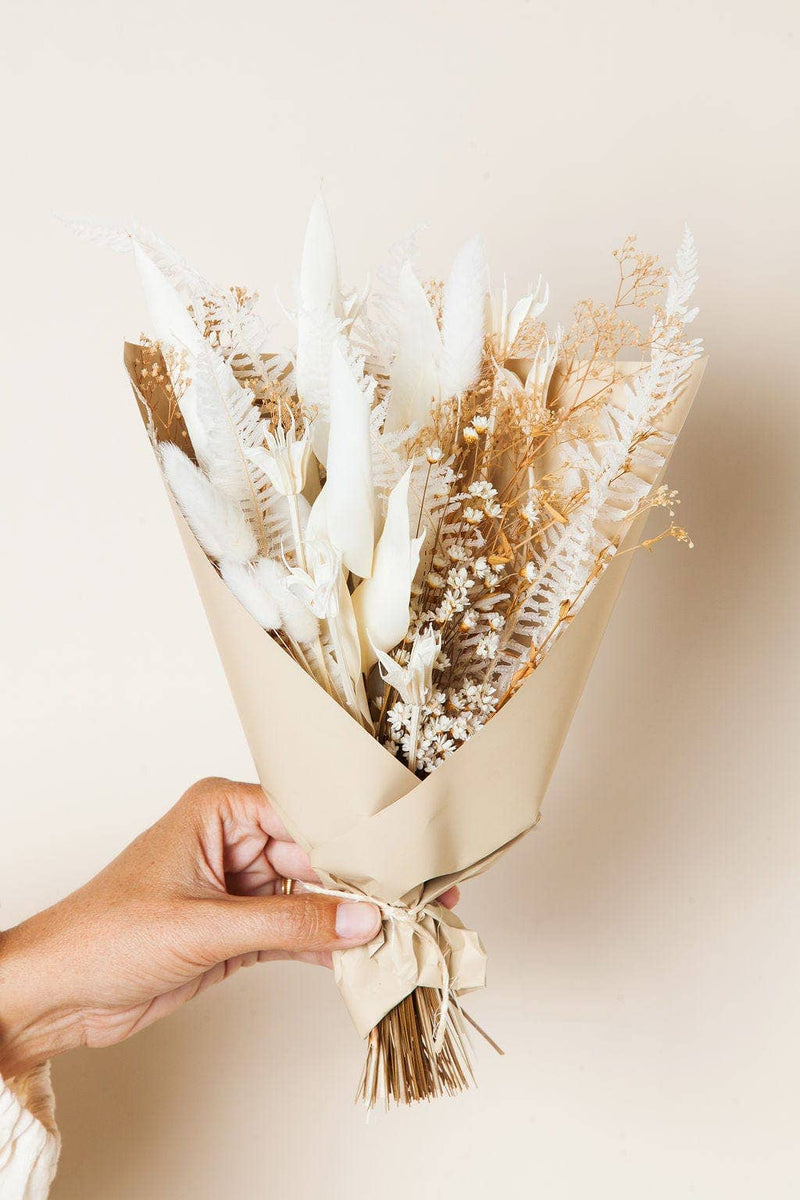 Idlewild Floral Co. - Snow Dried Flower Bouquet: A hand holds a small, beige-toned bouquet of dried flowers and grasses, wrapped in kraft paper and tied with twine.