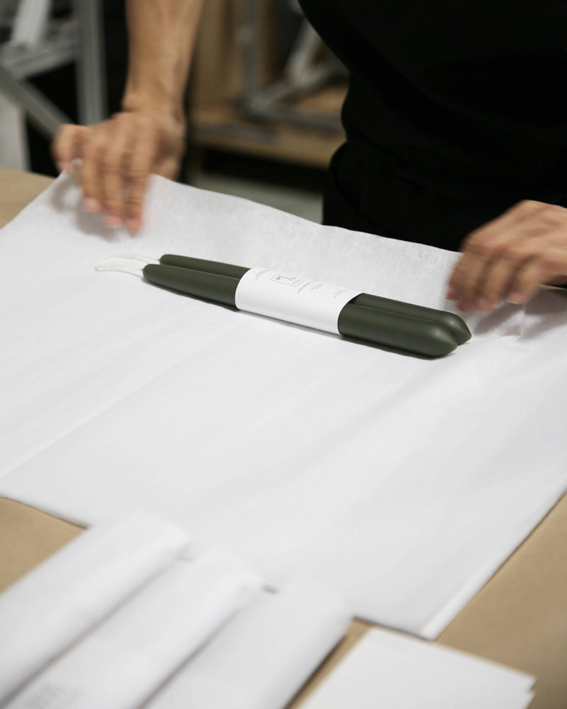 Mo&Co Home - Forest Green Dipped Beeswax Taper Candles, handcrafted, 14-inch, lie wrapped on white paper, hands folding for eco-friendly packaging.