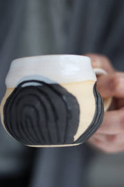 Hand holding the Gravesco Pottery Transformation Mug Petite, showcasing its cream and matte black design, symbolizing feminine strength and transformation in handcrafted elegance.