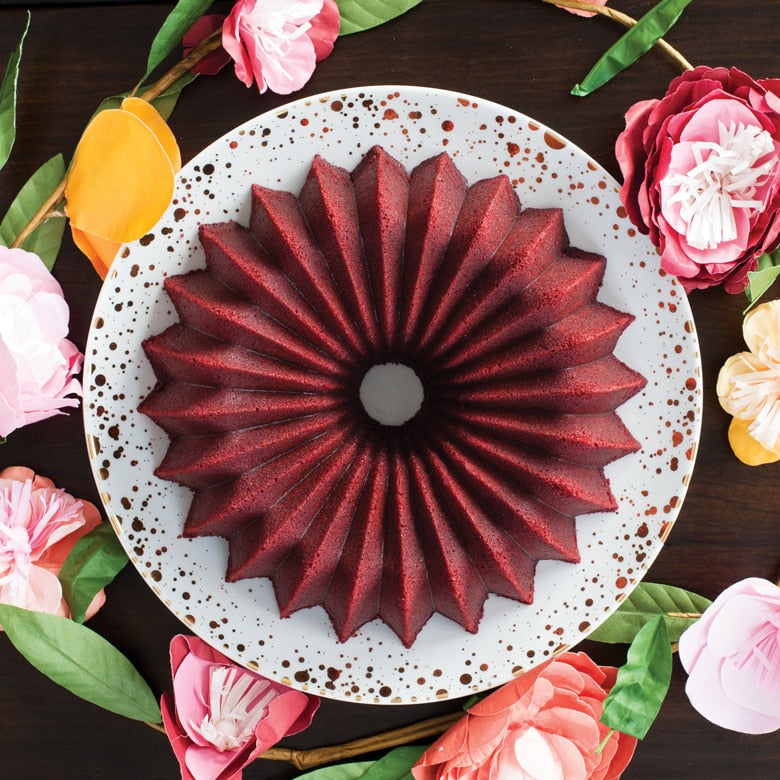 Nordic Ware | Brilliance Bundt® Pan featuring a red bundt cake on a plate, surrounded by flowers, showcasing its elegant design and nonstick surface.
