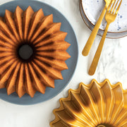 Nordic Ware | Brilliance 10-Cup Bundt pan showcasing a beautifully baked bundt cake on a plate, accompanied by elegant gold utensils, highlighting its exquisite design and quality.