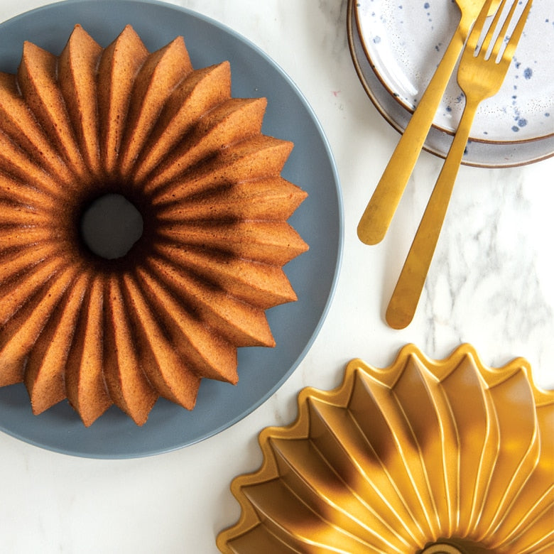 Nordic Ware | Brilliance 10-Cup Bundt pan showcasing a beautifully baked bundt cake on a plate, accompanied by elegant gold utensils, highlighting its exquisite design and quality.