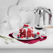 Dualit Hand Mixer in polished chrome stands beside red-velvet pancakes with cream and berries, showcasing its sleek design amidst a vibrant breakfast setting.