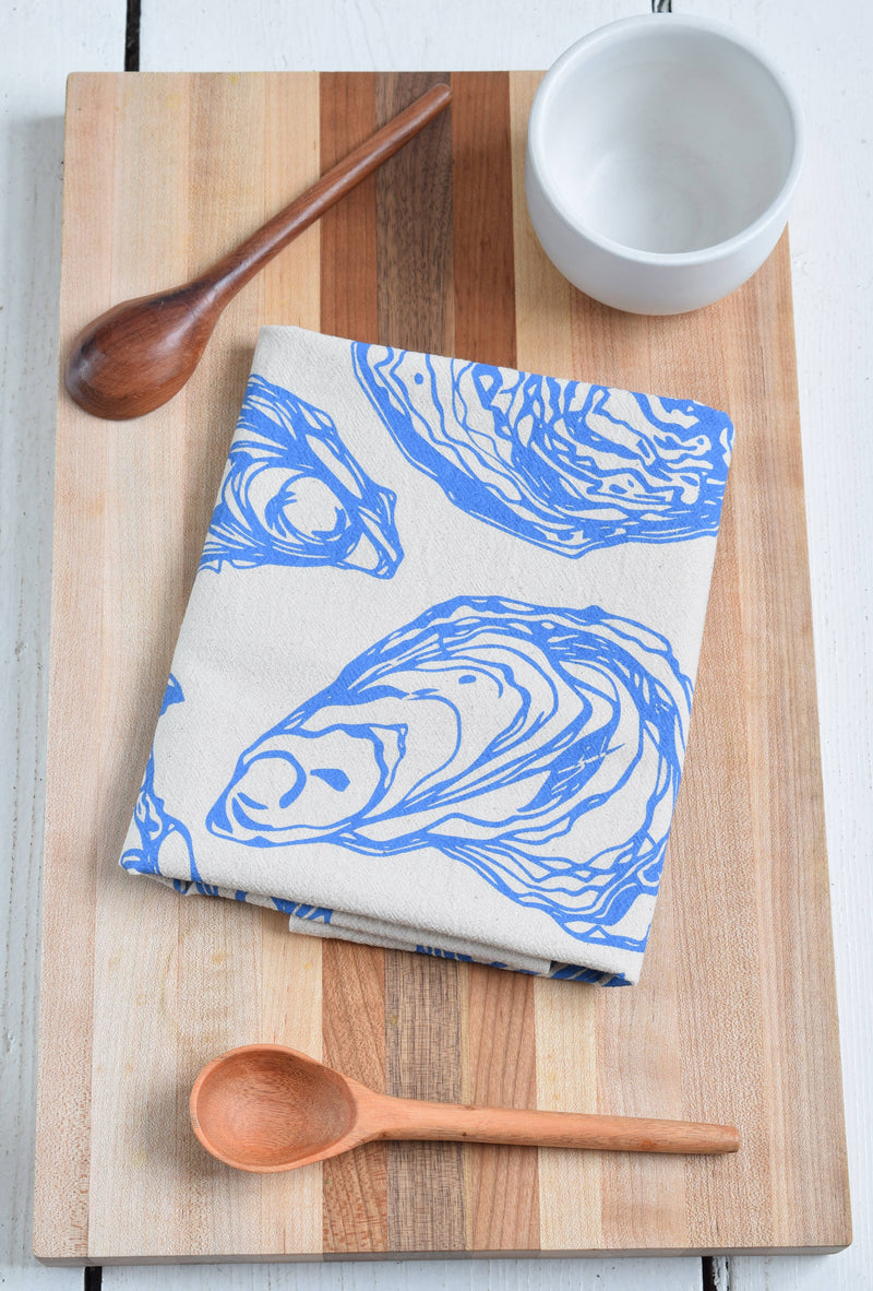 Hearth and Harrow - Oyster Tea Towel in Blue-violet - Organic Cotton displayed on a cutting board with a wooden spoon, showcasing its intricate sea shell design.