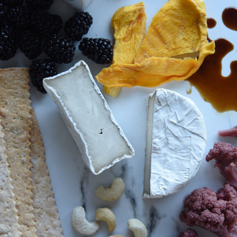Rebel Cheese - Cave-Aged Truffle Brie Plant Based, 5.4 oz, on a marble surface with blackberries and a block of cheese.