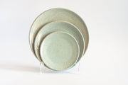 RachaelPots - Dinner Plate: Light Blue, a set of three speckled ceramic plates in varying sizes, displayed on a clear stand, showcasing their flat design with slightly upturned edges.
