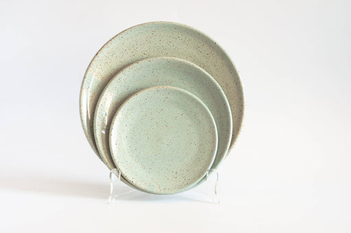 RachaelPots - Dinner Plate: Light Blue, a set of three speckled ceramic plates in varying sizes, displayed on a clear stand, showcasing their flat design with slightly upturned edges.