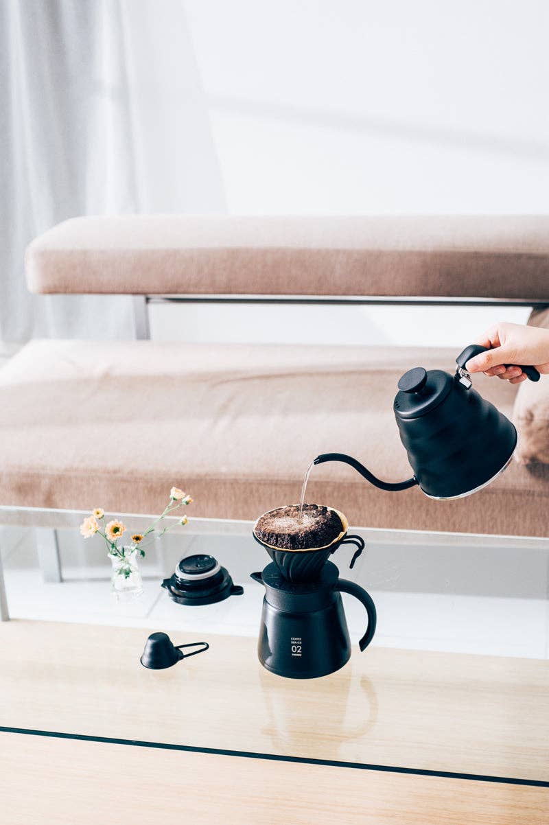 HARIO USA - V60 Metal Dripper: Silver in action, showcasing precision coffee brewing on a glass table with a gooseneck kettle and stylish accessories.