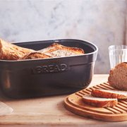 Emile Henry | Bread Box, Burgundy on a light wood countertop with sliced rustic loaf on a wooden board, showcasing its elegant ceramic design and multifunctional lid.