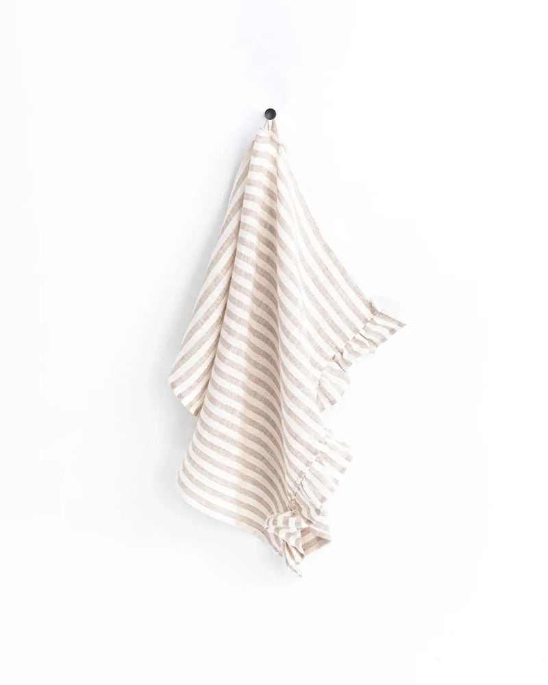 Linen tea towel with ruffle trim hanging on a hook; durable and absorbent, perfect for any kitchen.
