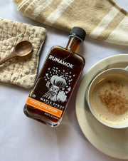Runamok - Pumpkin Spice Infused Organic Maple Syrup bottle with a spiced latte, wooden spoon, and kitchen textiles on a table, perfect for autumn recipes.