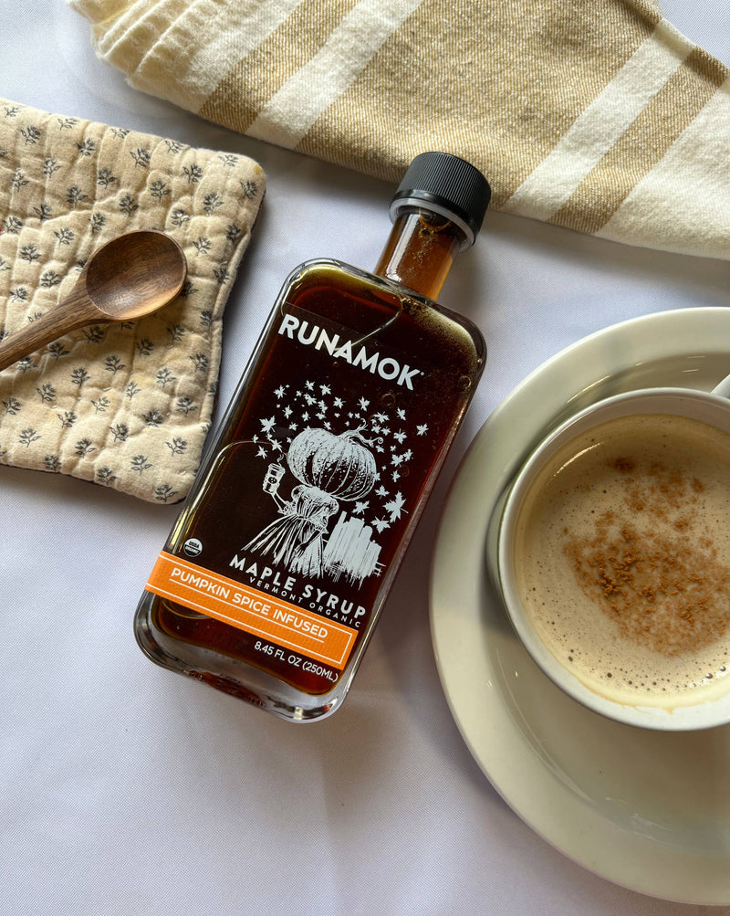 Runamok - Pumpkin Spice Infused Organic Maple Syrup bottle with a spiced latte, wooden spoon, and kitchen textiles on a table, perfect for autumn recipes.