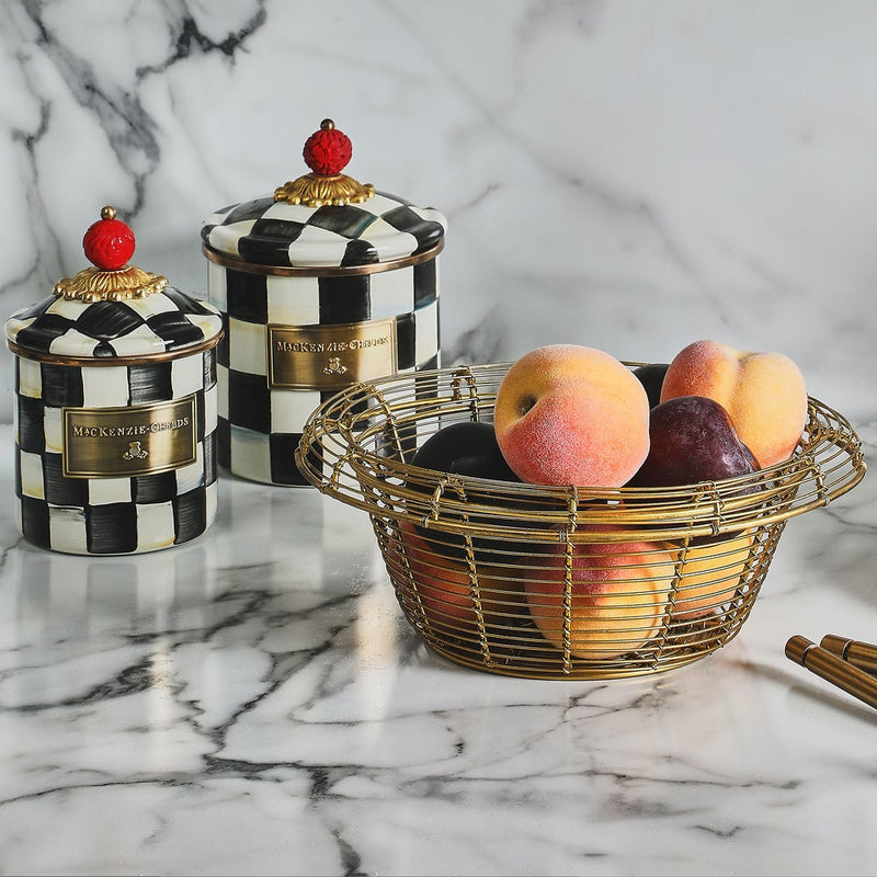 MacKenzie-Childs Courtly Check Small Canister featuring a black-and-white checkerboard design with a red-knob lid, paired with a gold-tone wire fruit basket holding peaches.