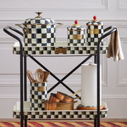 Two-tier black metal kitchen cart displaying a MacKenzie Childs | Courtly Check Large Canister set, with wooden utensils, copper pans, and a kitchen towel.