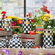 MacKenzie Childs Courtly Check Large Canister set amid vibrant garden flowers, showcasing hand-painted checks and cinnabar knobs on a rustic tabletop.