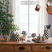 MacKenzie Childs | Courtly Check Large Canister on a rustic table, open to reveal cookies, surrounded by matching checkered kitchen accessories and a potted plant.