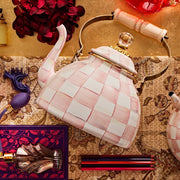 MacKenzie Childs | Rosy Check Tea Kettle, 3 QT: Porcelain kettle with pink-and-white checks, brass accents, wooden handle, surrounded by hydrangeas, perfume atomizer, and colored pencils.