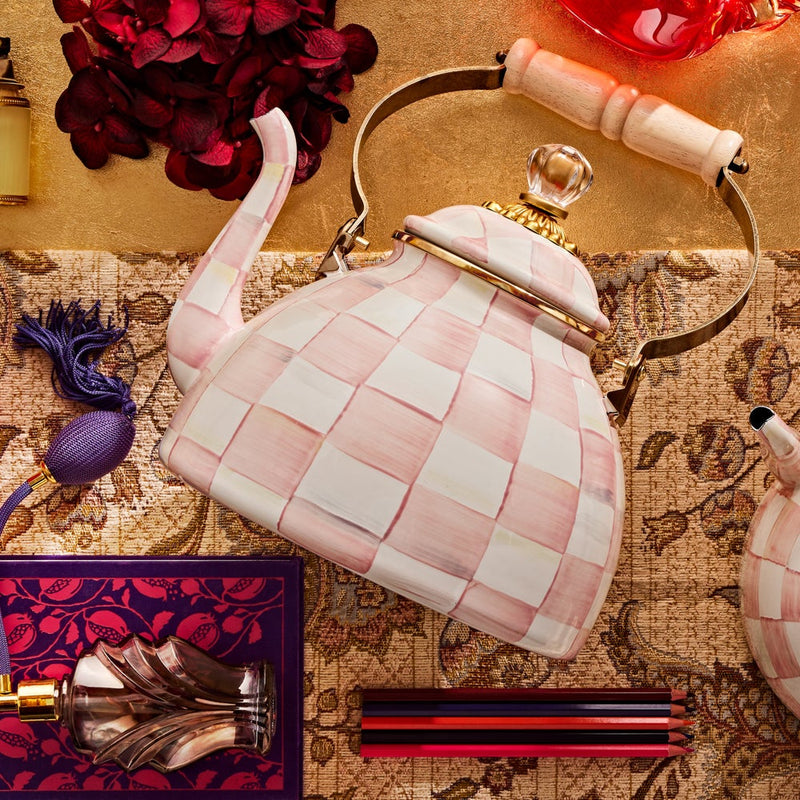 MacKenzie Childs | Rosy Check Tea Kettle, 3 QT: Porcelain kettle with pink-and-white checks, brass accents, wooden handle, surrounded by hydrangeas, perfume atomizer, and colored pencils.