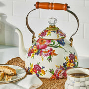 MacKenzie-Childs White Flower Market Tea Kettle with floral design, wooden handle, sits on placemat beside biscotti and matching mug in an Athens Cooks kitchen setting.