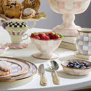MacKenzie-Childs Rosy Check Ice Cream Dish with hand-painted checker pattern, filled with strawberries, surrounded by matching tableware including a pedestal bowl, tiered stand, and gold-tone utensils.
