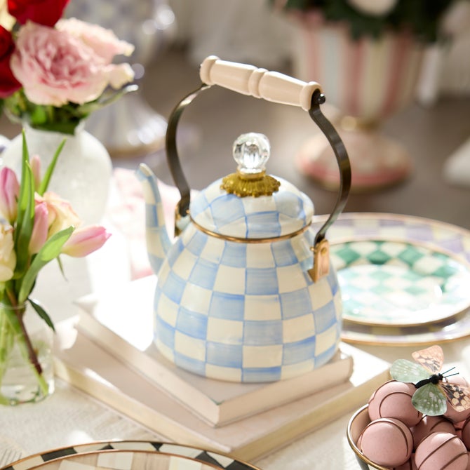 MacKenzie-Childs Sky Check Tea Kettle, 2 Qt, with a blue-and-white checkered pattern, sits on books amidst pink flowers and pastel tableware.