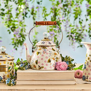 MacKenzie Childs Wildflowers Pink Tea Kettle, 2 QT., with polka dots and daisies, wood handle, glass knob, atop books, surrounded by blooms.