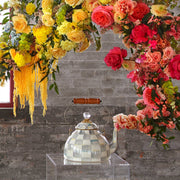 MacKenzie Childs | Sterling Check Tea Kettle, 2 QT., displayed on an acrylic pedestal, features hand-painted checks, wood handle, and glass knob, surrounded by warm-hued flowers.