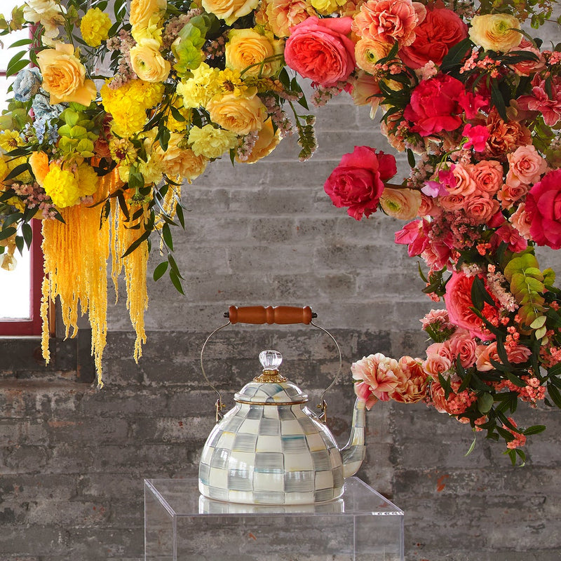 MacKenzie Childs | Sterling Check Tea Kettle, 2 QT., displayed on an acrylic pedestal, features hand-painted checks, wood handle, and glass knob, surrounded by warm-hued flowers.