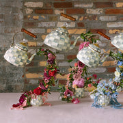 MacKenzie Childs | Sterling Check Tea Kettle, 2 QT, pouring vibrant flowers into matching teacups, creating a whimsical, floating floral display against a brick backdrop.