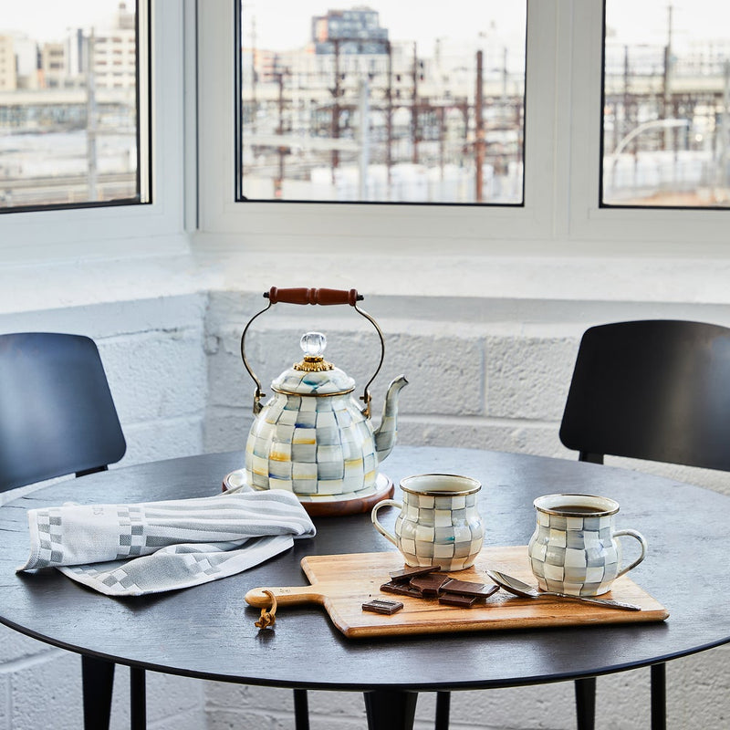 MacKenzie Childs | Sterling Check Tea Kettle, 2 QT., on sunlit table, features hand-painted checks, wooden handle, with matching mugs, chocolate, and tea towel nearby, in cozy kitchen setting.